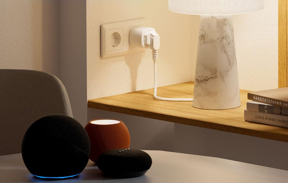 Τρία έξυπνα ηχεία Amazon Echo σε μαύρο, πορτοκαλί και μαύρο χρώμα τοποθετημένα σε ένα τραπέζι μπροστά από μια έξυπνη πρίζα συνδεδεμένη σε μια πρίζα ρεύματος και μια λάμπα σε ένα ξύλινο ράφι