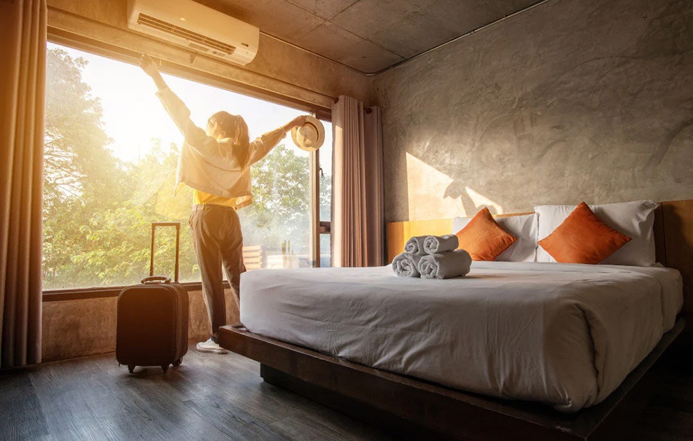 Kobieta przy oknie w hotelowym pokoju z dużym łóżkiem i klimatyzacją nad głową.