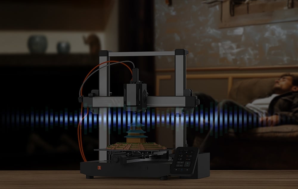 Drukarka Anycubic Kobra 3 V2, w tle widać mężczyznę, który odpoczywa na kanapie.