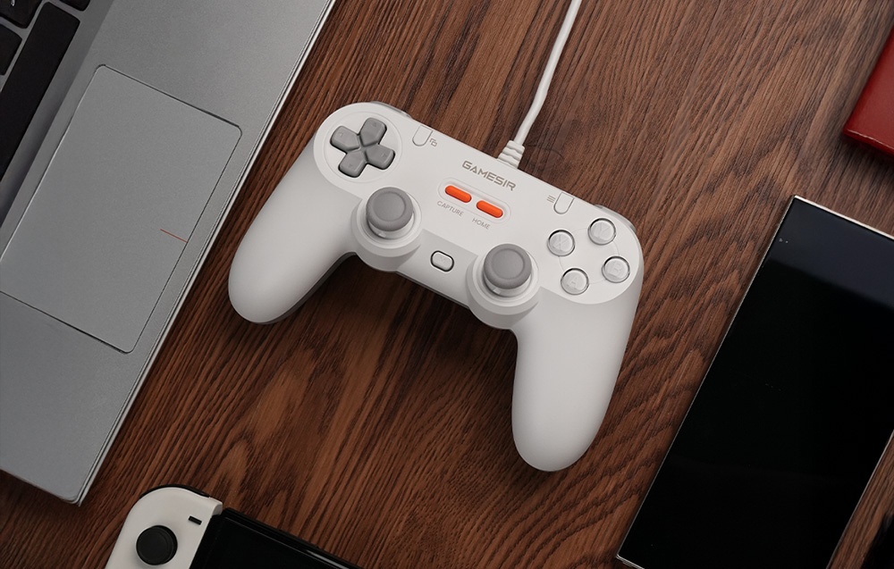 Λευκό χειριστήριο GameSir συνδεδεμένο με καλώδιο, τοποθετημένο σε ξύλινο τραπέζι δίπλα σε φορητό υπολογιστή και smartphones.