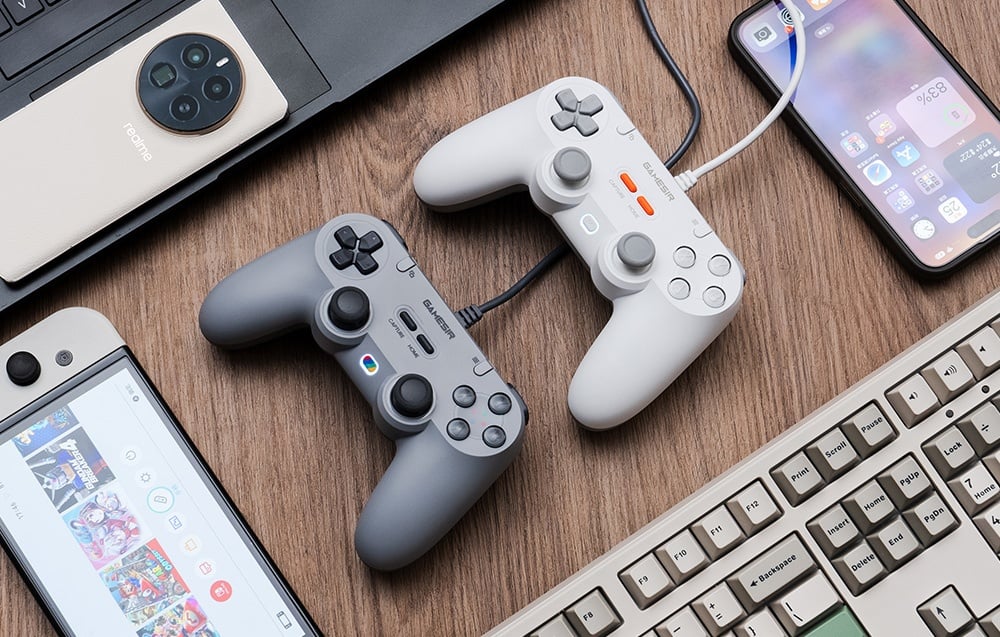 Ένα λευκό και ένα γκρι χειριστήριο GameSir με καλώδιο στις συσκευές, που βρίσκονται ανάμεσα στα ηλεκτρονικά είδη πάνω στο ξύλινο γραφείο.