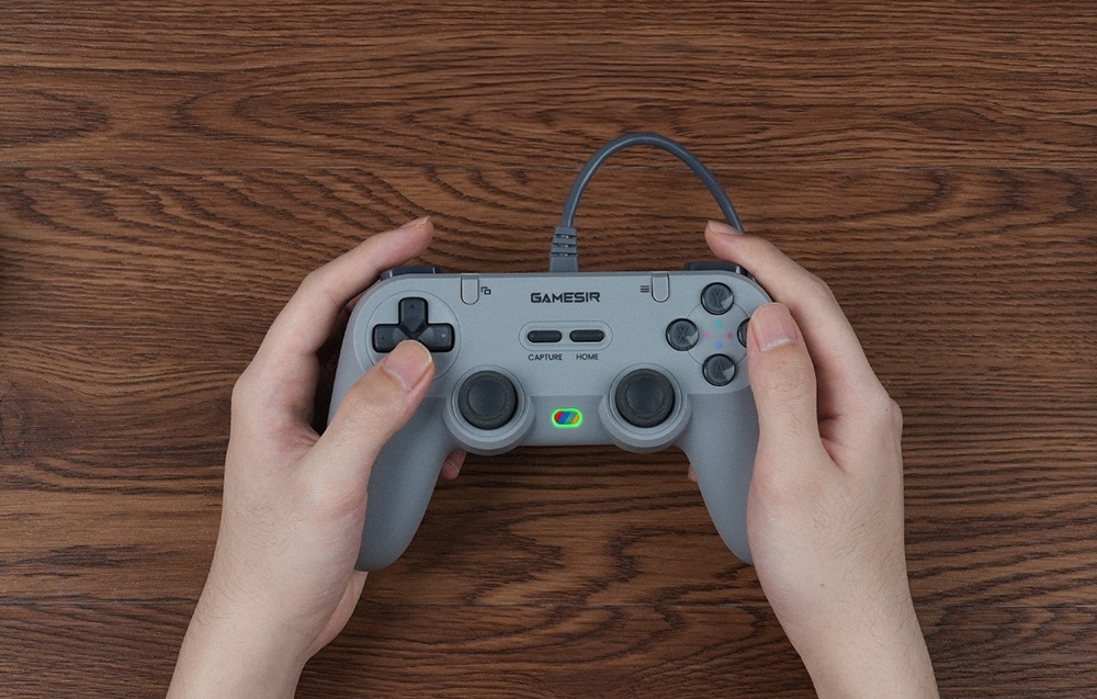 Ένα άτομο κρατάει ένα γκρι χειριστήριο GameSir με τα δύο χέρια πάνω από ένα ξύλινο τραπέζι.