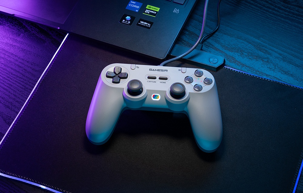 Ένα γκρίζο χειριστήριο GameSir ξαπλωμένο σε ένα μαξιλάρι μπροστά από ένα φορητό υπολογιστή, φωτισμένο από μπλε-ιώδες φως.