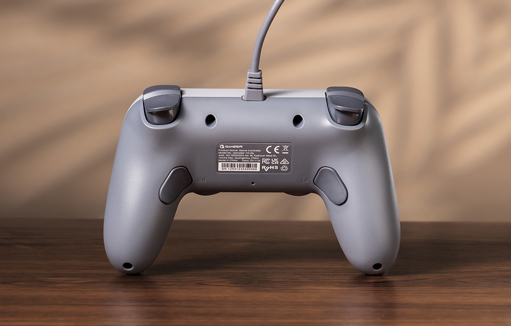 Το πίσω μέρος του γκρίζου χειριστηρίου GameSir με τα κουμπιά, την ετικέτα και το καλώδιο ορατά πάνω στο ξύλινο τραπέζι.
