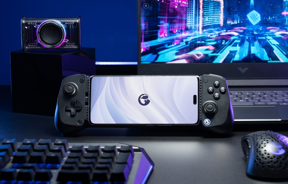 Ένα smartphone τοποθετημένο σε ένα μαύρο χειριστήριο GameSir τοποθετημένο σε ένα γραφείο μπροστά από ένα φορητό υπολογιστή και αξεσουάρ παιχνιδιών.