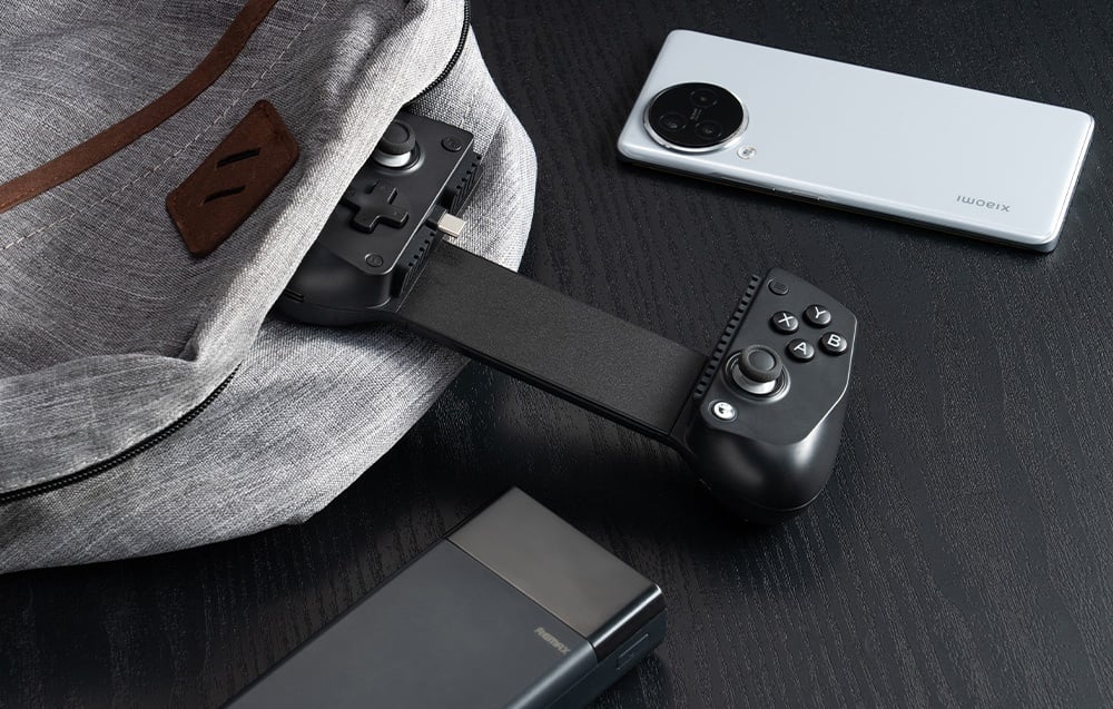 Μαύρο χειριστήριο GameSir που προεξέχει μερικώς από σακίδιο πλάτης, δίπλα σε smartphone και powerbank σε μαύρο τραπέζι.
