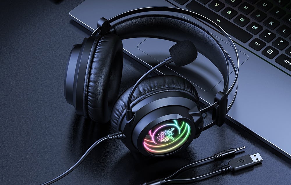 Ακουστικά με οπίσθιο φωτισμό RGB ξαπλωμένα δίπλα σε φορητό υπολογιστή με καλώδια USB και jack σε σκοτεινό γραφείο