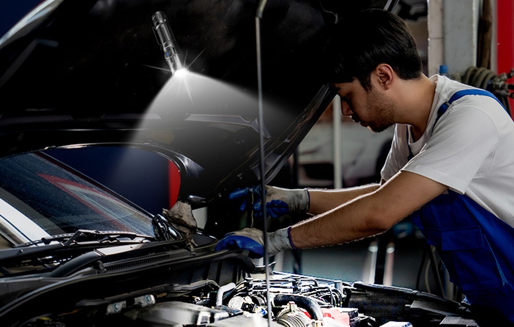 Μηχανικός με μπλε στολή φτιάχνει τον κινητήρα αυτοκινήτου χρησιμοποιώντας φακό για φωτισμό
