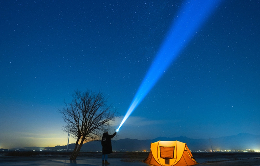 Σιλουέτα ατόμου με φακό που εκπέμπει μπλε δέσμη φωτός προς τον ουρανό δίπλα σε μια πορτοκαλί σκηνή