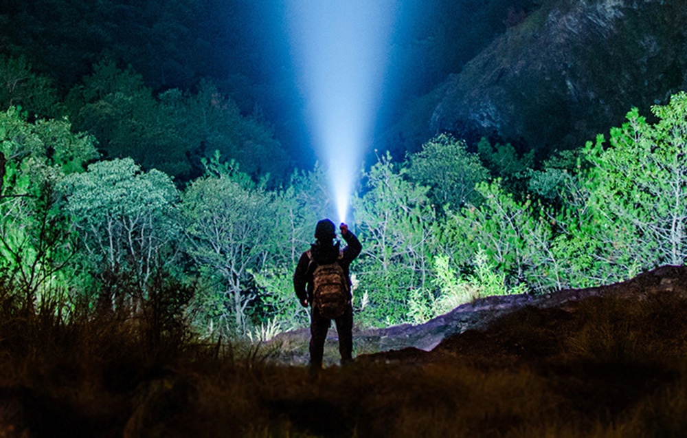 Άτομο με σακίδιο πλάτης κατευθύνει μπλε δέσμη φωτός από φακό επάνω ανάμεσα σε πράσινα δέντρα τη νύχτα