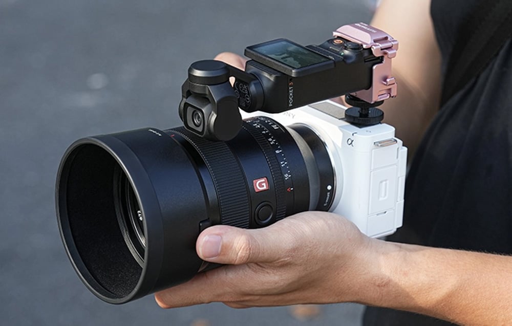 Μια φωτογραφική μηχανή DJI Pocket σε ροζ βάση στήριξης τοποθετημένη πάνω σε μια φωτογραφική μηχανή Sony με μεγάλο φακό, που κρατείται στα χέρια