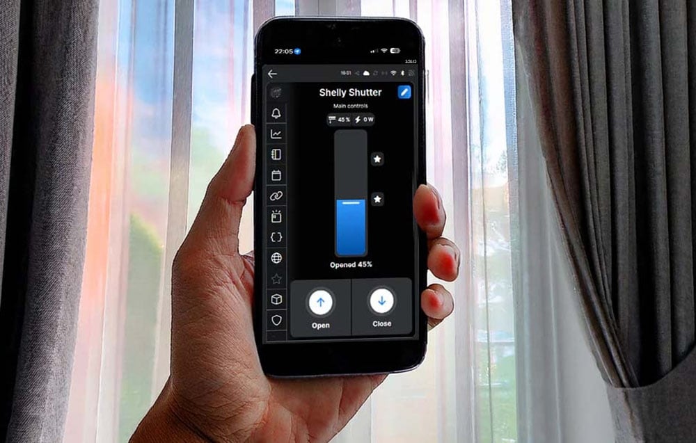 Smartphone με την εφαρμογή Shelly Shutter στο χέρι, που δείχνει τον έλεγχο των περσίδων παραθύρων με φόντο ημιδιαφανείς κουρτίνες