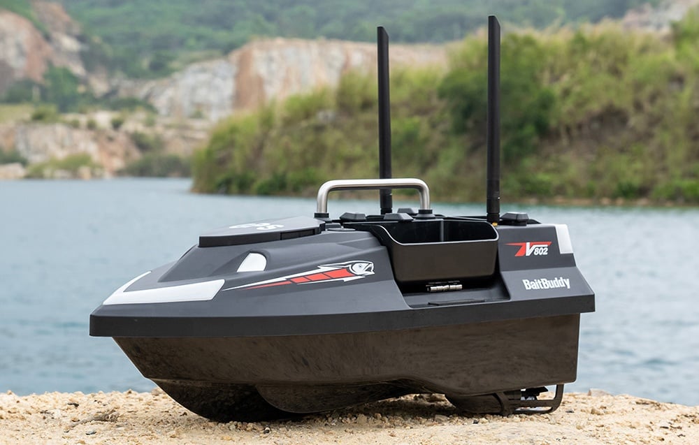 BaitBuddy V602 RC σκάφος δόλωμα με δύο κεραίες που στέκεται σε μια αμμώδη όχθη λίμνης με βουνά στο φόντο.