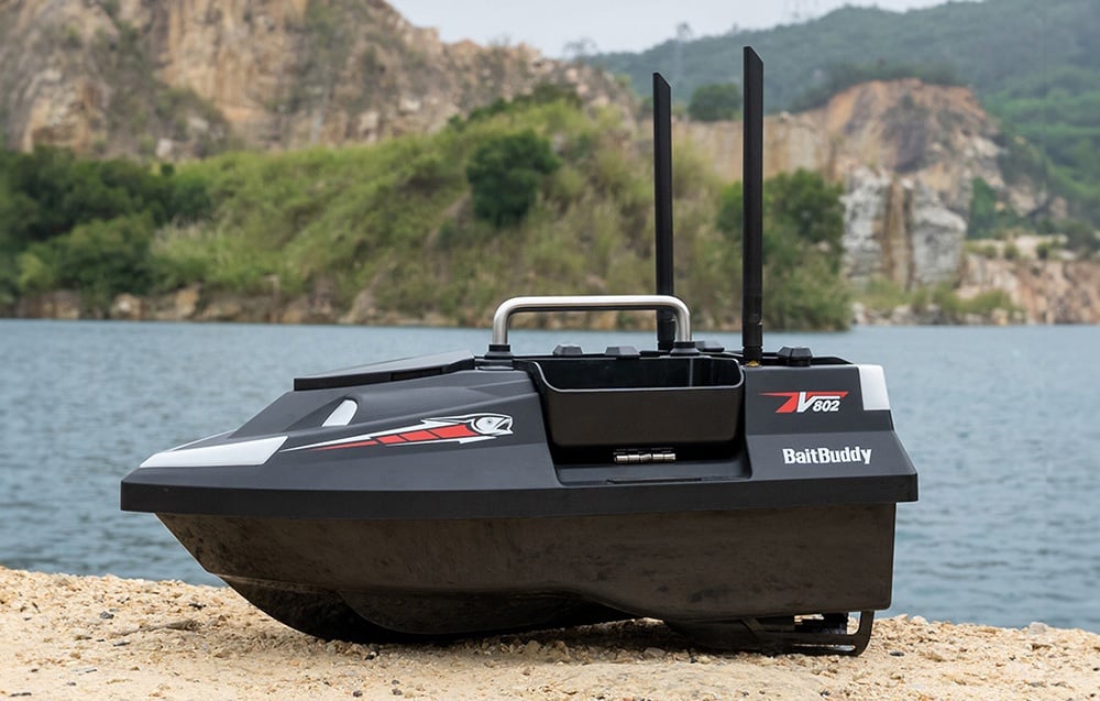 RC BaitBuddy V602 σκάφος δόλωμα με δύο κεραίες που στέκεται στην όχθη της λίμνης με τα βουνά στο φόντο.