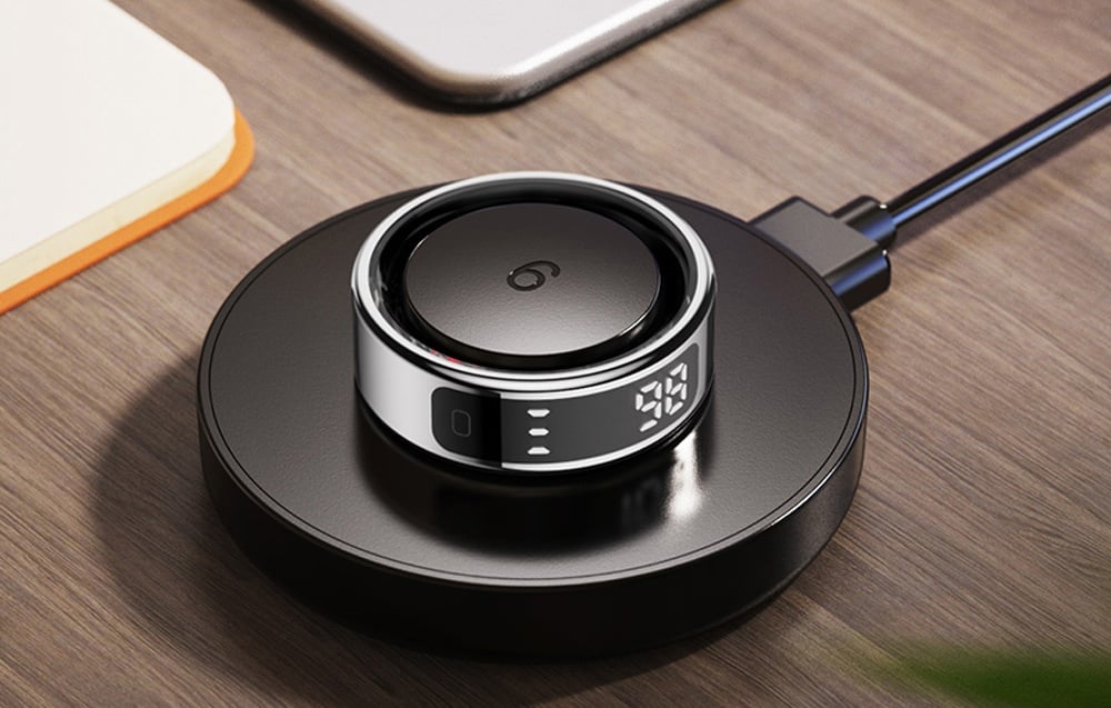 Smartring με οθόνη σε βάση φόρτισης σε ξύλινο τραπέζι.