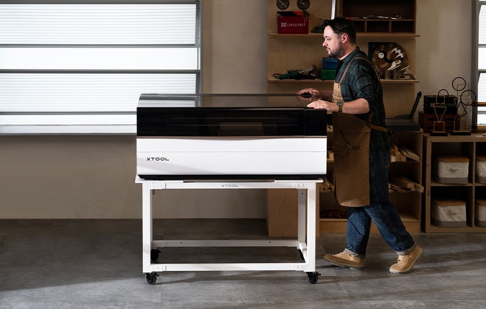 Ένας άνδρας με ποδιά στέκεται δίπλα σε ένα xTool laser engraver πάνω σε ένα λευκό καρότσι στο εργαστήριο