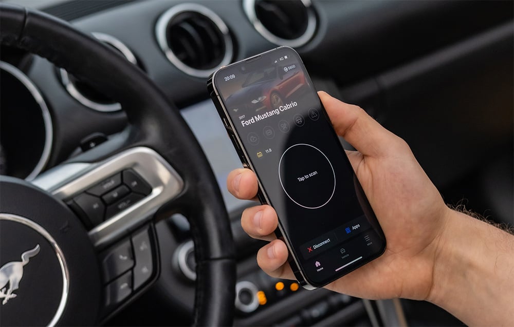 Χέρι που κρατά smartphone με την εφαρμογή Carly που εμφανίζει δεδομένα Ford Mustang Convertible μέσα στο αυτοκίνητο