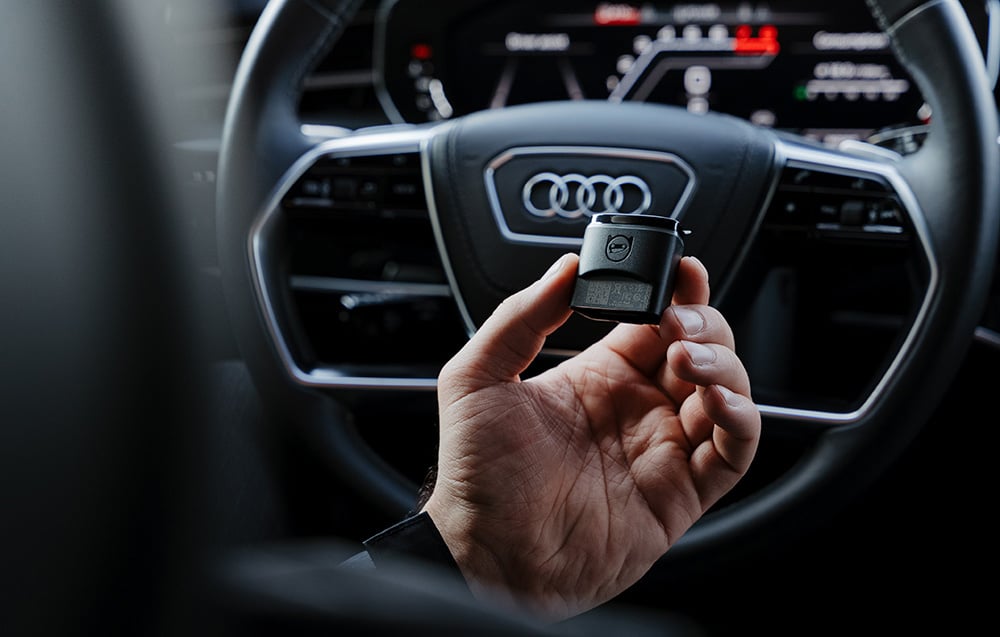 Διαγνωστικός προσαρμογέας Carly OBD2 που κρατείται μπροστά από το τιμόνι με το λογότυπο της Audi στο ψηφιακό πιλοτήριο
