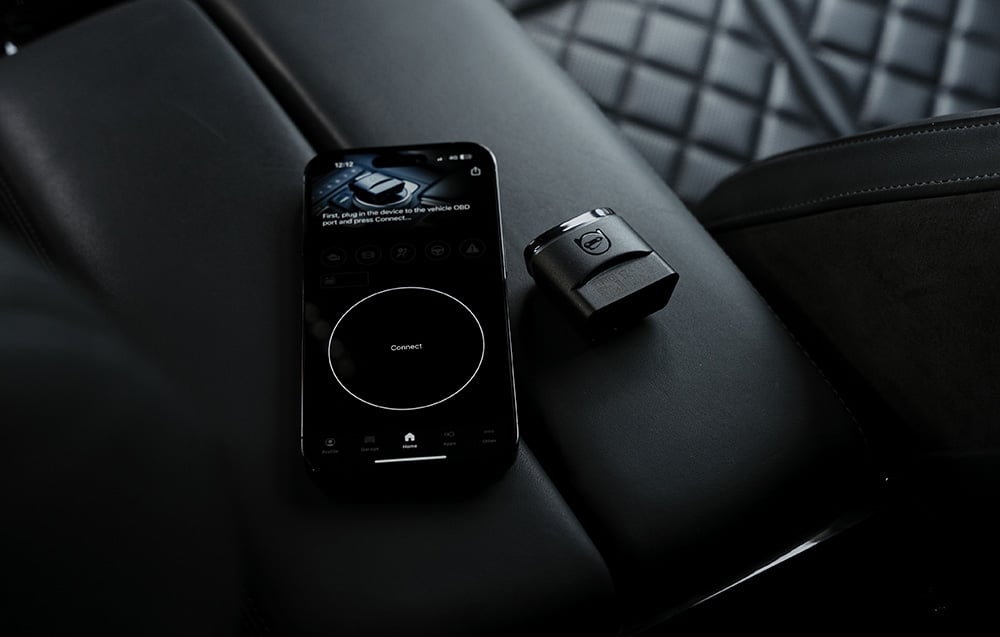 Smartphone με την εφαρμογή Carly δίπλα στον προσαρμογέα OBD2 σε μαύρο καπιτονέ κάθισμα αυτοκινήτου