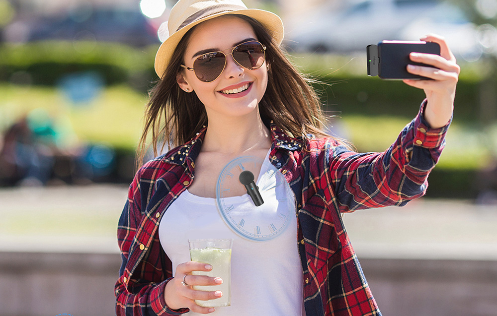 Μια γυναίκα σε εξωτερικό χώρο βγάζει selfie με ένα smartphone με ένα μικρόφωνο καρφωμένο σε αυτό και κρατάει ένα ποτό σε ένα διαφανές ποτήρι.