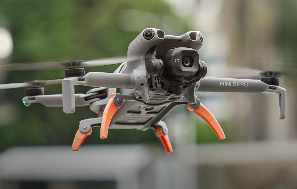 DJI Mini 5 Pro ιπτάμενο drone με απλωμένα πορτοκαλί πόδια, που αιωρείται στον αέρα