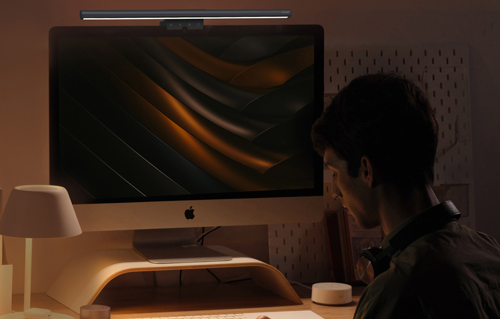Ένας άνδρας κάθεται μπροστά από έναν iMac με λαμπτήρα τοποθετημένο στο πάνω μέρος της οθόνης, σε ένα σκοτεινό δωμάτιο