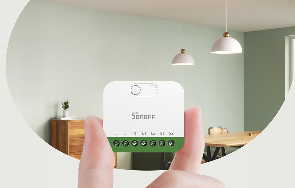 Λευκός έξυπνος διακόπτης Sonoff MINI-2GS που κρατιέται στα χέρια και δείχνει τον ακροδέκτη με τις ενδείξεις σύνδεσης L, N, L1, L2, S1, S2, με φόντο ένα μοντέρνο σαλόνι με κρεμαστά φώτα