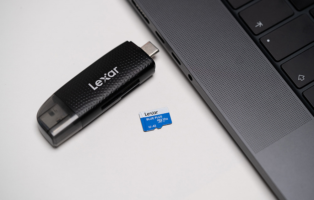 Κάρτα μνήμης Lexar Blue Plus microSDXC πάνω σε λευκό τραπέζι δίπλα σε μαύρο αναγνώστη καρτών USB-C και στην άκρη ενός γκρι φορητού υπολογιστή.