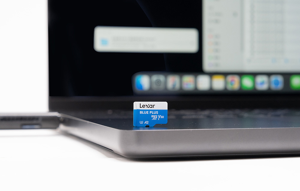 Κοντινό πλάνο της κάρτας Lexar Blue Plus microSD που στέκεται όρθια πάνω σε θήκη φορητού υπολογιστή, με θολή οθόνη που εμφανίζει τη διεπαφή του συστήματος στο παρασκήνιο.