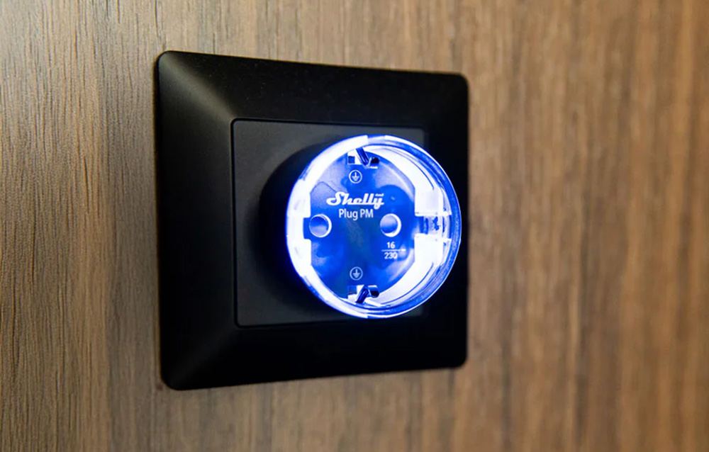 Έξυπνη πρίζα Shelly Plug PM με μπλε οπίσθιο φωτισμό LED, τοποθετημένη σε μαύρο πλαίσιο σε ξύλινο τοίχο.