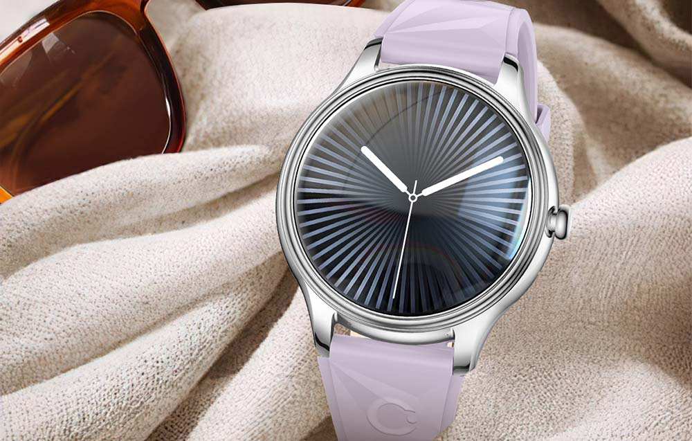 Ένα ασημένιο smartwatch με μοβ γεωμετρικό λουράκι και σκούρο ριγέ καντράν, τοποθετημένο πάνω σε ανοιχτόχρωμο ύφασμα δίπλα σε γυαλιά ηλίου.
