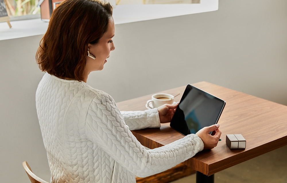 Μια γυναίκα με λευκό πουλόβερ κρατάει ένα tablet και φοράει ακουστικό μετάφρασης