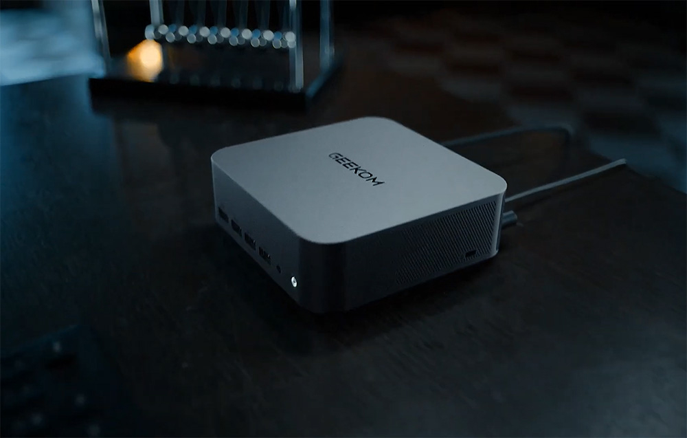 Ένα γκρι MINI PC Geekom με καλώδιο συνδεδεμένο σε ένα μαύρο γραφείο