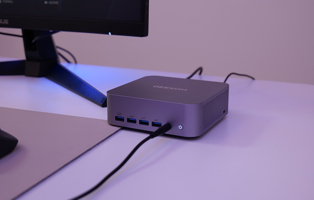Ένα γκρι Geekom MINI PC με ένα καλώδιο συνδεδεμένο σε αυτό βρίσκεται πάνω σε ένα γραφείο δίπλα σε μια οθόνη σε ένα δωμάτιο με μοβ φωτισμό
