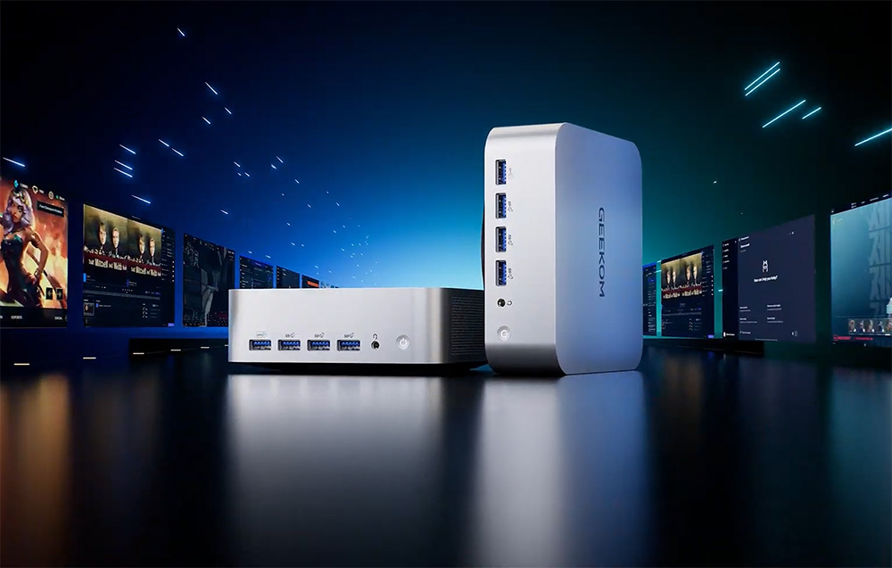 2 γκρι Geekom Mini PC σε μαύρο πάγκο, οθόνες στα πλάγια