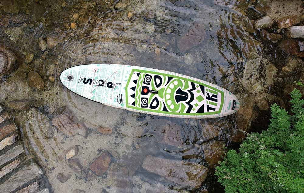 Μια πολύχρωμη σανίδα σέρφινγκ Funwater βρίσκεται στο νερό, περιτριγυρισμένη από βράχους και φυτά
