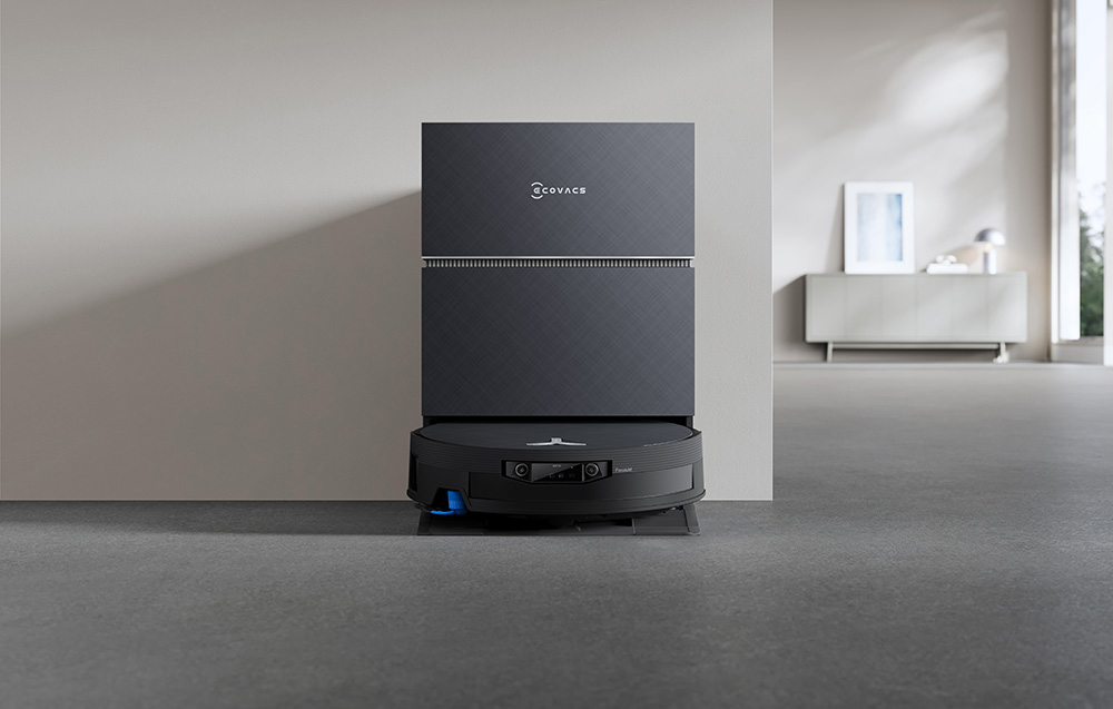 Το ρομπότ καθαρισμού Ecovacs αγκυροβολημένο σε έναν κομψό, μαύρο σταθμό εκκένωσης μπροστά από έναν ανοιχτόχρωμο τοίχο.