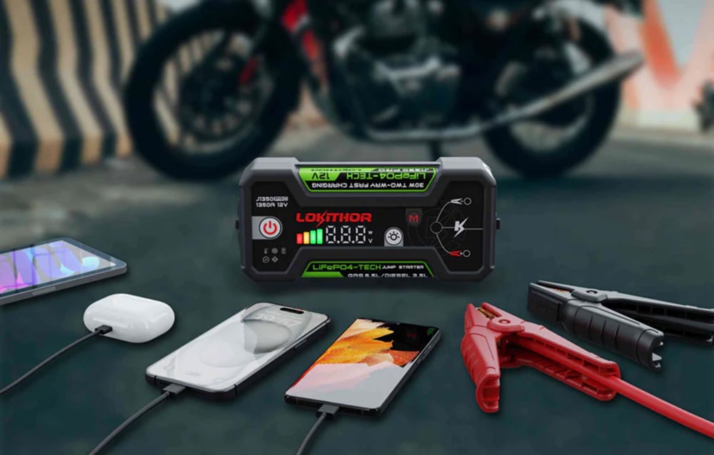 Το LOKITHOR jump starter πάνω στον άσφαλτο φορτίζει smartphone και ακουστικά δίπλα σε μια μοτοσικλέτα.
