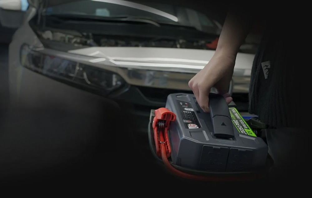 Ένα φορητό Lokithor jump starter κρατημένο από τη λαβή με το καπό του αυτοκινήτου ανοιχτό το βράδυ.
