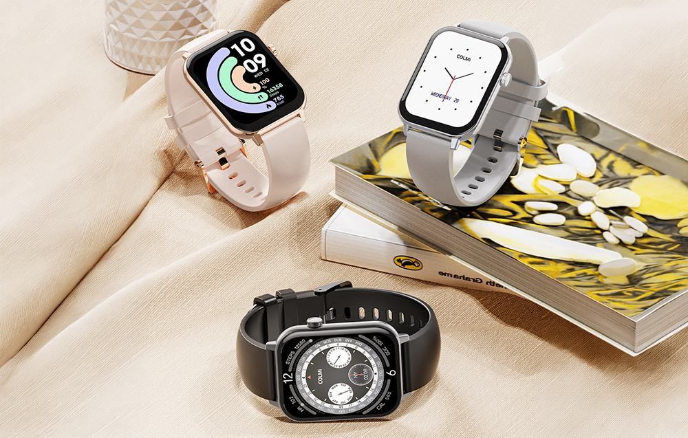 Τρία smartwatch Colmi (ροζ, γκρι, μαύρο) σε μια κομψή διάταξη πάνω σε ύφασμα δίπλα σε βιβλία