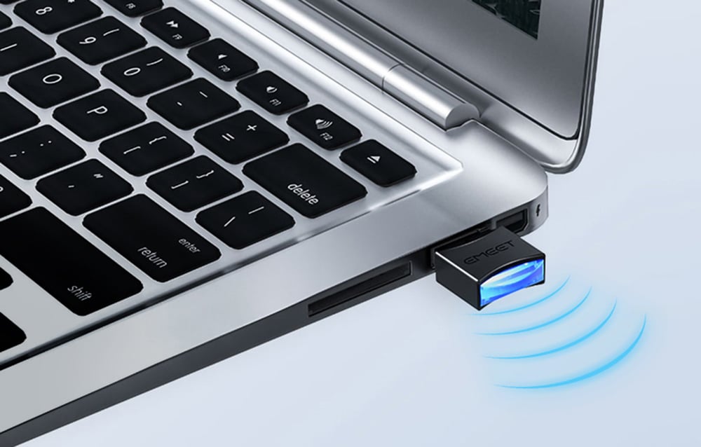 Κοντινό πλάνο ενός μικρού, μαύρου δέκτη USB με λογότυπο και μπλε οπίσθιο φωτισμό, συνδεδεμένου στην πλευρική θύρα ενός ασημένιου φορητού υπολογιστή
