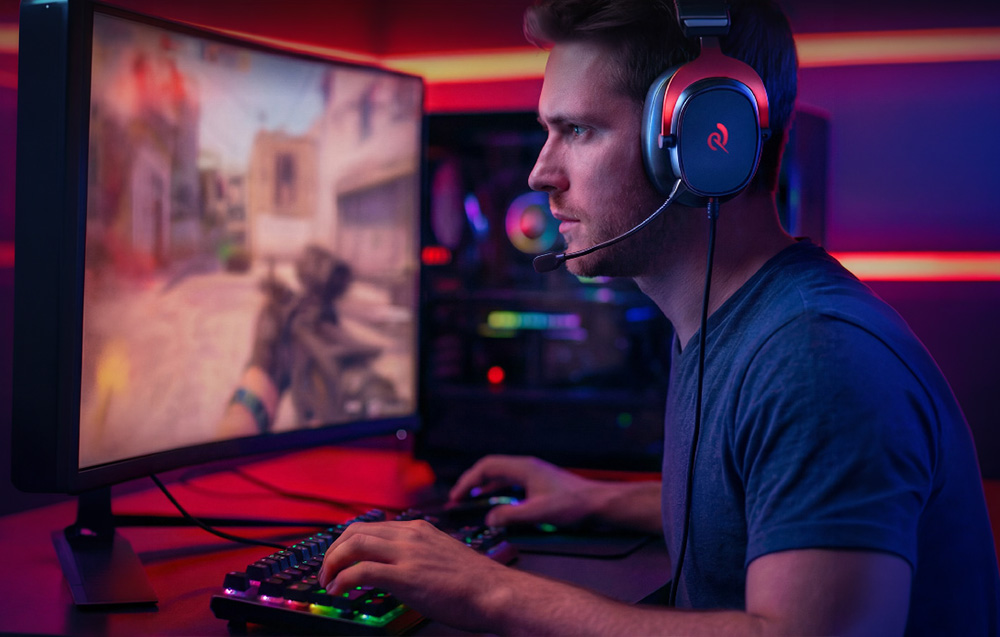 Ένας άνδρας που φοράει ακουστικά gaming και παίζει ένα FPS shooter σε μια καμπύλη οθόνη σε ένα δωμάτιο με κόκκινο και μπλε φωτισμό