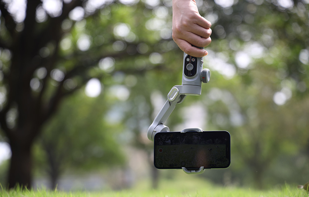 Ένα χέρι κρατά ένα gimbal με ένα smartphone στραμμένο προς τα κάτω ενώ τραβάει βίντεο σε εξωτερικό χώρο.
