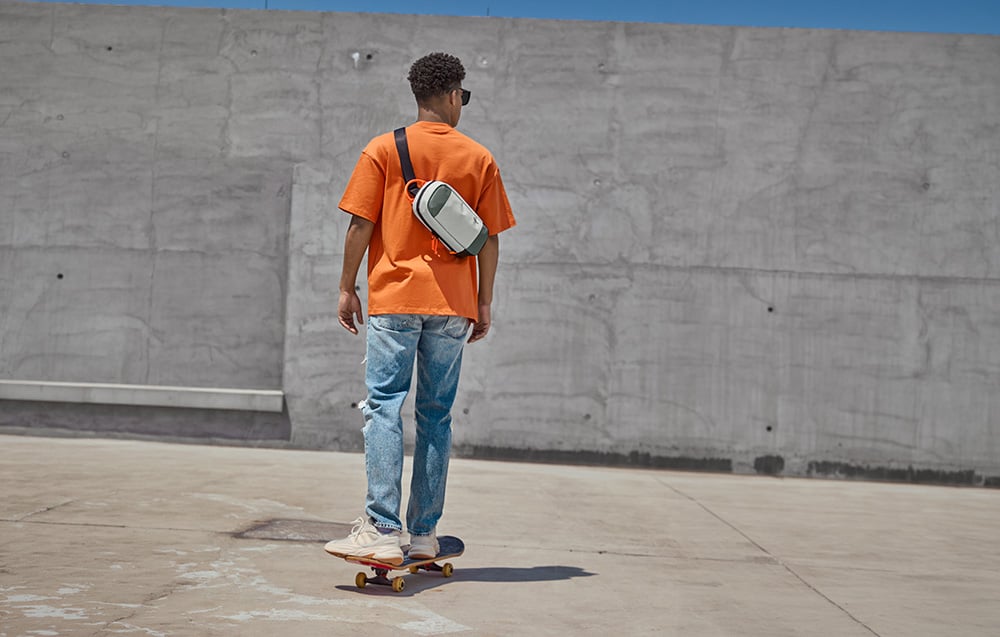 Ένας άνδρας που φαίνεται από πίσω να κάνει skateboard· κρατά μια γκρι τσάντα ώμου· ένας τοίχος από μπετόν στο βάθος