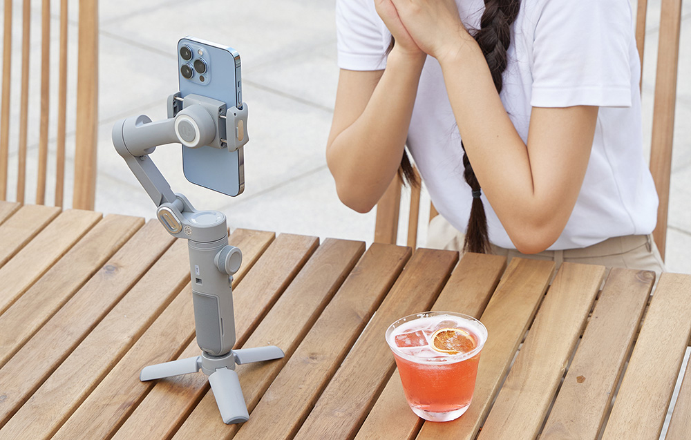 Ένα smartphone σε ένα gimbal τοποθετημένο σε τρίποδο καταγράφει ένα πλάνο δίπλα σε ένα τραπέζι με ένα ποτό.