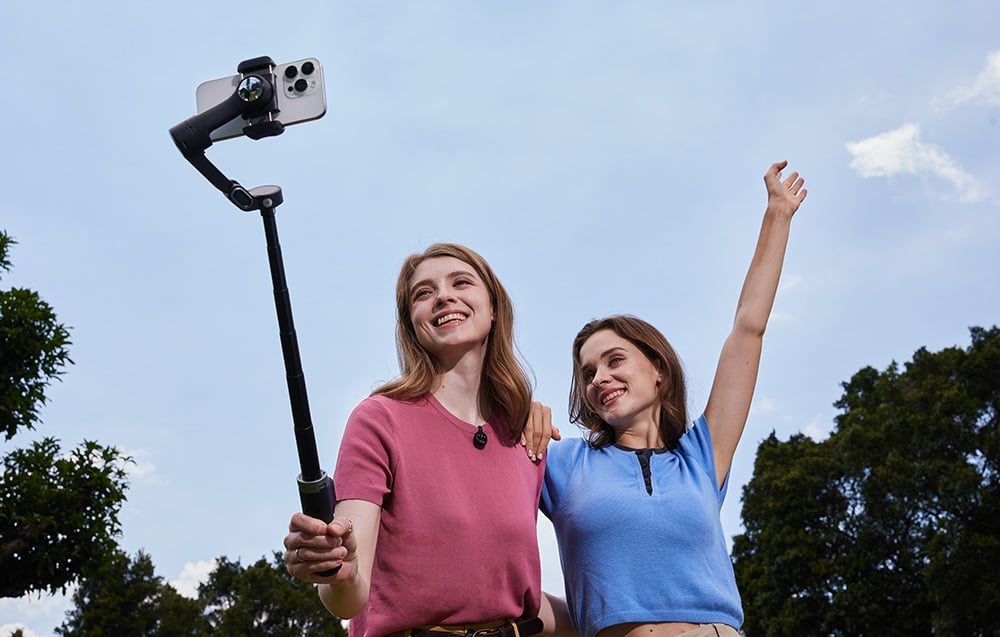 Δύο γυναίκες τραβούν μια selfie μαζί σε εξωτερικό χώρο, χρησιμοποιώντας ένα gimbal με βραχίονα επέκτασης.