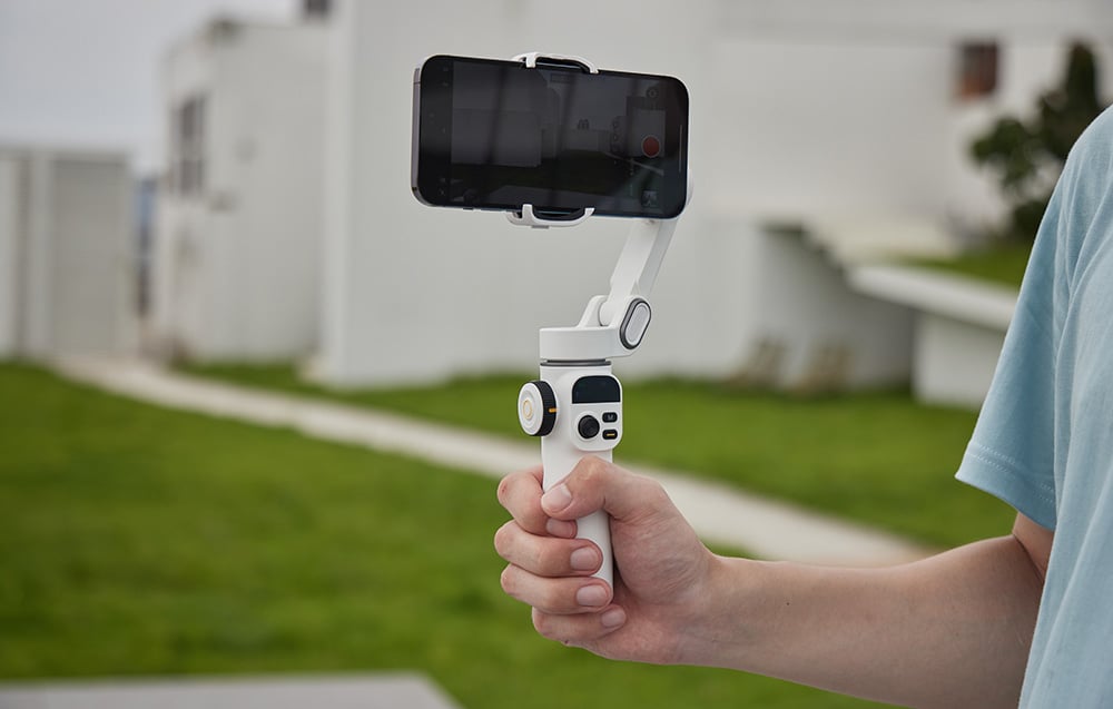 Ένα χέρι που κρατά οριζόντια ένα λευκό gimbal με ένα smartphone ενώ πραγματοποιεί εγγραφή σε εξωτερικό χώρο.