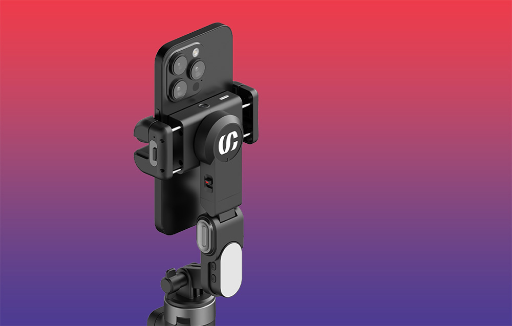 Κοντινό πλάνο της βάσης στήριξης του gimbal με ένα smartphone συνδεδεμένο και τη μονάδα σταθεροποίησης πάνω σε φόντο με κόκκινο-ιώδες ντεγκραντέ.