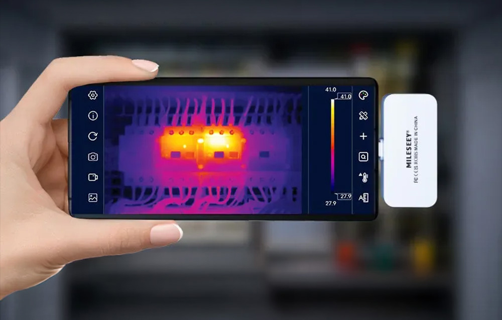 Ένα smartphone με θερμική κάμερα στο χέρι δείχνει σημεία υψηλής θερμοκρασίας σε έναν πίνακα ελέγχου.