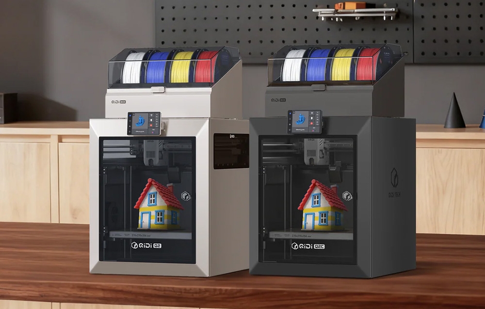 Δύο εκτυπωτές 3D QIDI με πολύχρωμα νήματα εκτυπώνουν μοντέλα μικρών σπιτιών πάνω σε ένα ξύλινο τραπέζι